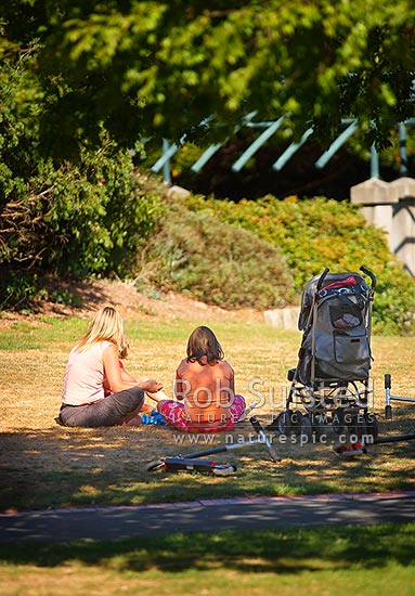 Family having picnic on lawn in sunny summer park. Solo parent, single mother, children with pram and toys, New Zealand (NZ)