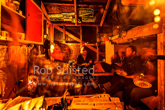 Musterers eating dinner by fire and candle light at the old Hundred Mile Hut. Early to bed, for a 4am start, Branches Station, Shotover Valley, Queenstown Lakes District, Otago Region, New Zealand (NZ)