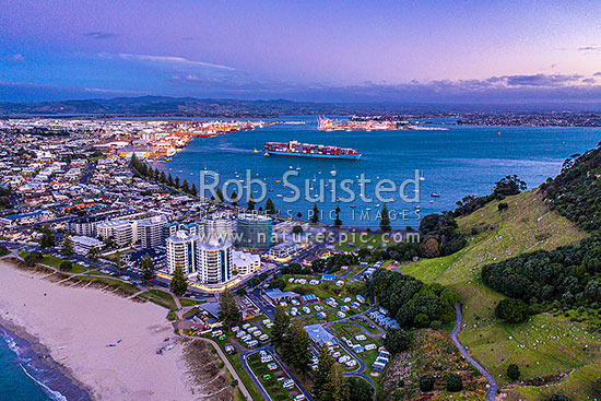 Mt Maunganui beach and township, with Tauranga Harbour and Port behind, with container ship departing. Aerial view at dusk, Mount Maunganui, Tauranga District, Bay of Plenty Region, New Zealand (NZ)