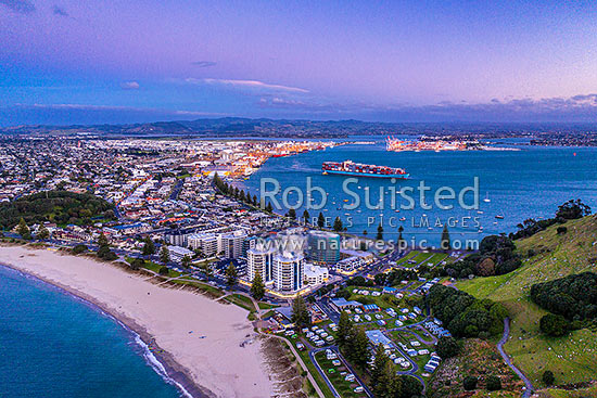 Mt Maunganui beach and township, with Tauranga Harbour and Port behind, with container ship departing. Aerial view at dusk, Mount Maunganui, Tauranga District, Bay of Plenty Region, New Zealand (NZ)